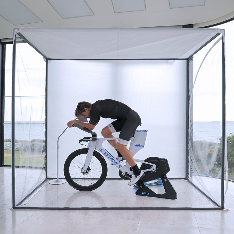 Cyclist in an aero tuck on a time-trial bike and Tacx trainer inside the clear-walled enclosure, sea view behind